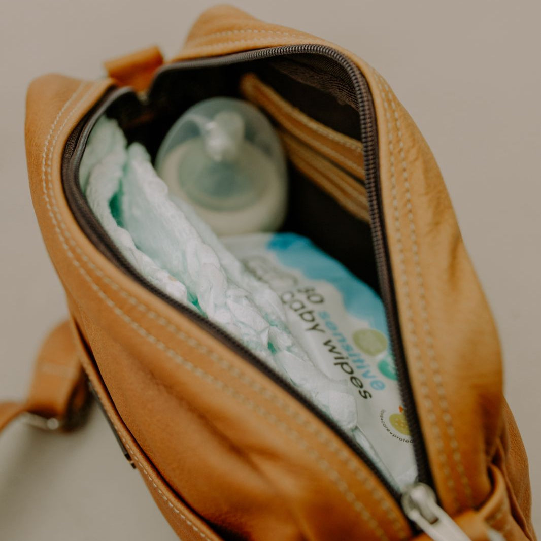 Open brown leather bag revealing baby items including diapers and a bottle on a light background